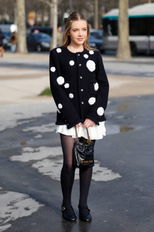 Guest wears black cardigan with white flowers decorations, white mini skirt, black tights, black Chanel bag, black Chanel mary jane shoes, outside...