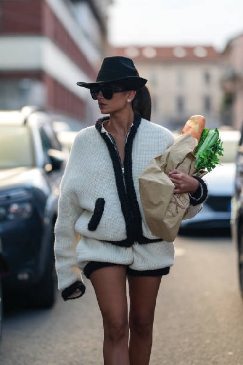 Guest wears black cowboy hat, black sunglasses, white oversized black lined fluffy jacket, white black line fluffy mini shorts, brown bag with bread...