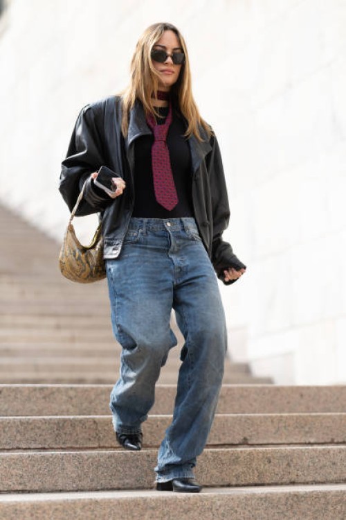 Guest wears black leather heeled boots, a yellow leather bag, brown sunglasses, a dark red printed tie, a black t-shirt, a dark brown leather bag and...