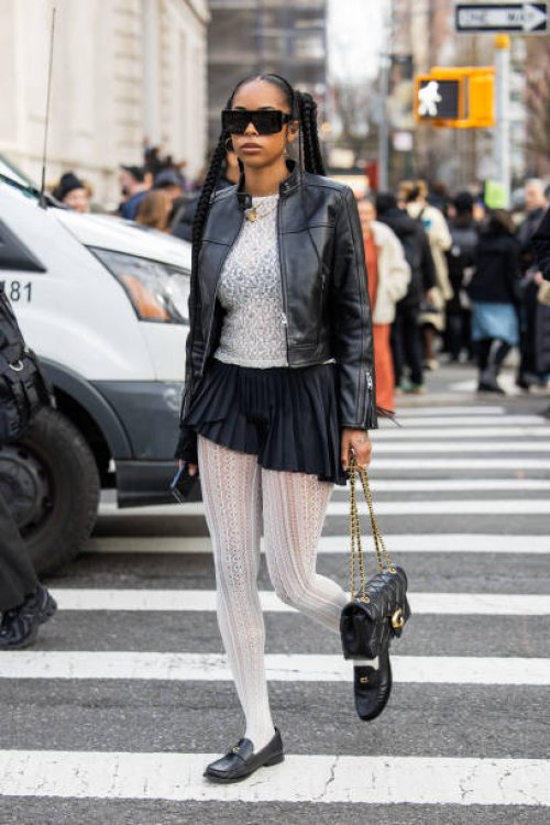 Guest wears black leather jacket, skirt, white laced tights outside Coach on February 12, 2024 in New York City.