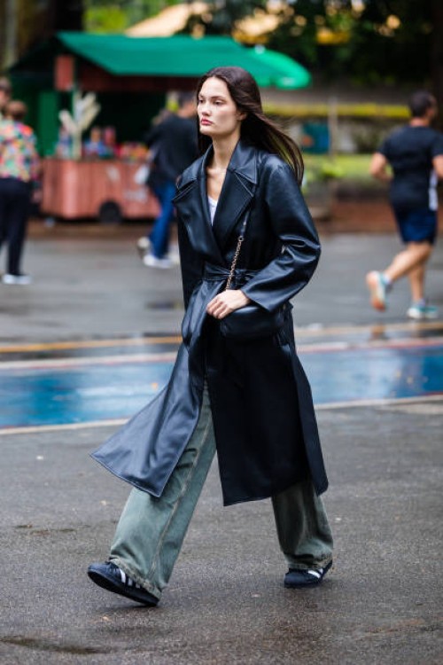 Guest wears black leather trench coat, white top, loose green wide-leg jeans, black sneakers, black crossbody bag with chain strap, minimalist style...