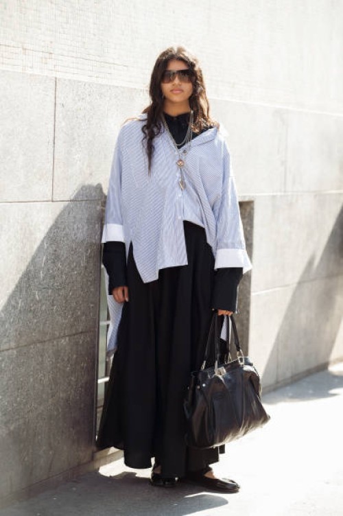Guest wears black long skirt, black turtle neck, blue and white oversized shirt, statement silver necklaces and sunglasses outside the Avavav fashion...