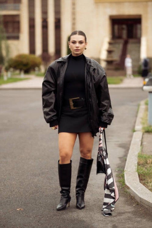 Guest wears black mini dress with a belt , leather jacket, long boots, black bag and a scarf wears during the 10th season of the Visa Fashion Week...