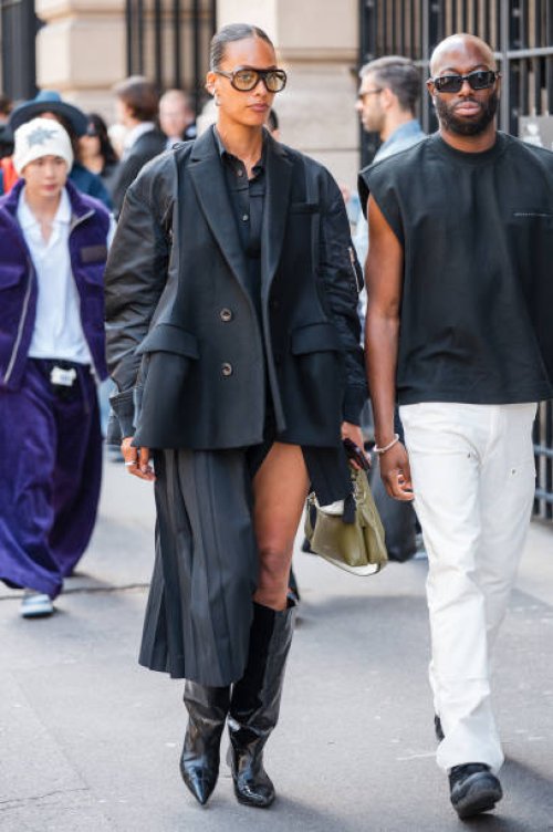 Guest wears black shirt, black jacket with blazer details, black pleated skirt, black boots, outside Sacai, during the Haute Menswear Spring/Summer...