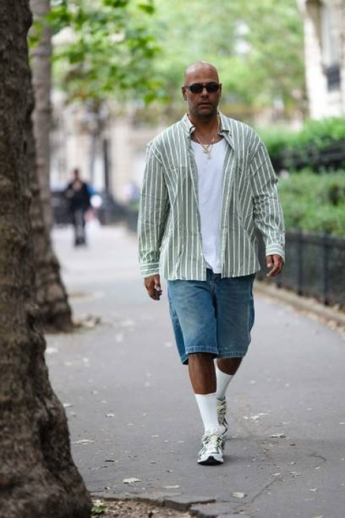 Guest wears black small sunglasses, earrings, a white t-shirt, a white and green striped print pattern shirt, gold chain pendant necklaces, blue...