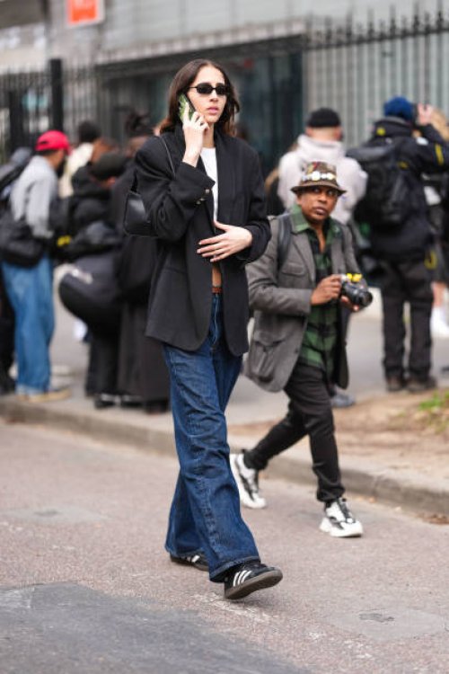 Guest wears black sunglasses, a black blazer jacket, a white top, a black leather bag, blue denim jeans / pants, black and white sneakers shoes,...