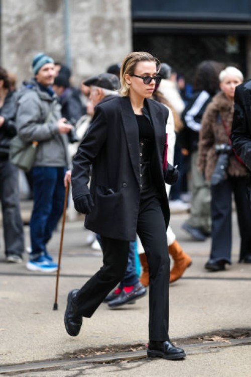 Guest wears black sunglasses, a black oversized blazer jacket, black pants, black gloves, black leather shoes, outside Dolce & Gabbana, during the...