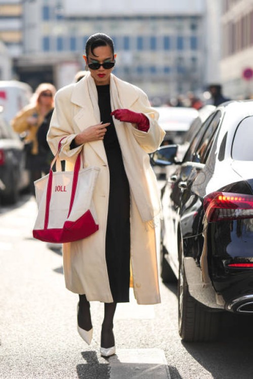 Guest wears black sunglasses, a black wool pullover, a red shiny leather high glove, black short legging pants, a white latte oversized long coat, a...