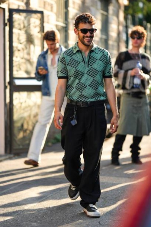 Guest wears black sunglasses, a green with embroidered black pattern short sleeves polo shirt, a black shiny leather belt, black large suit pants,...