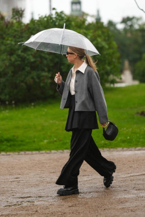 Guest wears black sunglasses, a white latte shirt, a gray blazer jacket, a black pleated / accordion short skirt, a black shiny leather handbag from...