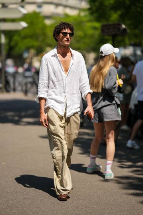 Guest wears black sunglasses, a white linen cotton shirt, beige large pants, dark brown shiny leather shoes , outside Hermes, during the Menswear...