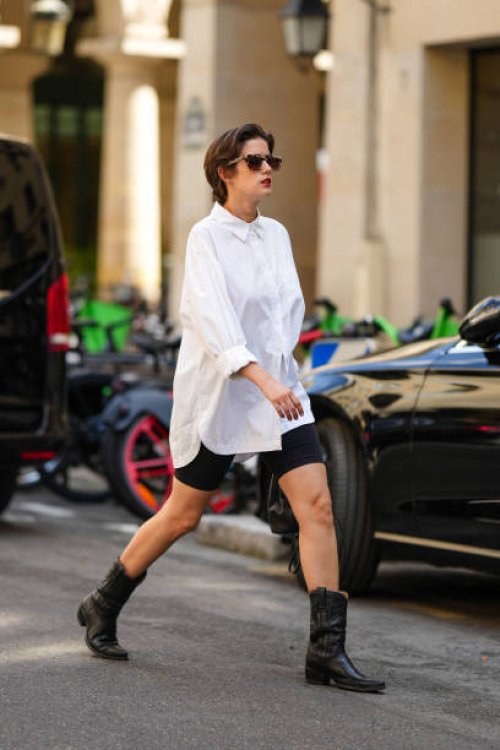 Guest wears black sunglasses, a white oversized shirt, black cyclist shorts, black shiny leather pointed western ankle boots , outside Fendi, during...