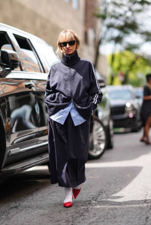 Guest wears black sunglasses, black high track suit jacket, light blue buttoned up long sleeve shirt, black midi skirt, white socks, shiny red...