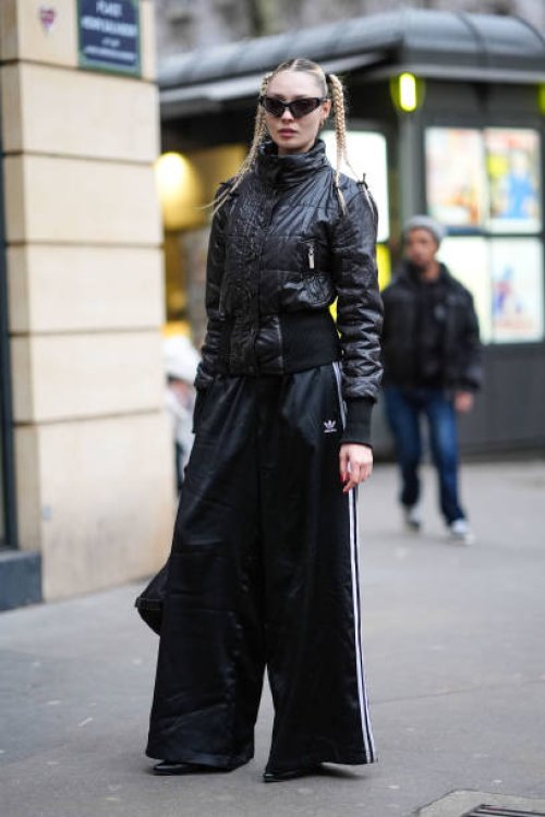 Guest wears black sunglasses, black leather turtleneck jacket, black oversized pants with white stripes, a black leather bag, outside Vetements,...
