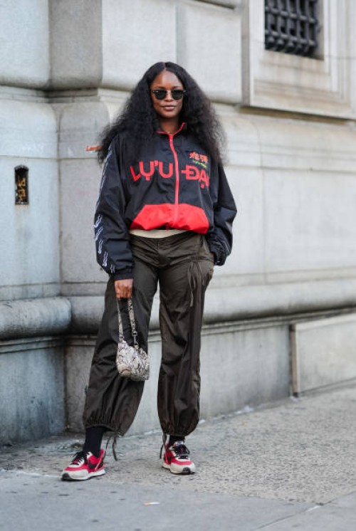 Guest wears black sunglasses, black red graphic oversized jacket, dark green baggy pants, light gray pattern/print leather bag, white socks, red...
