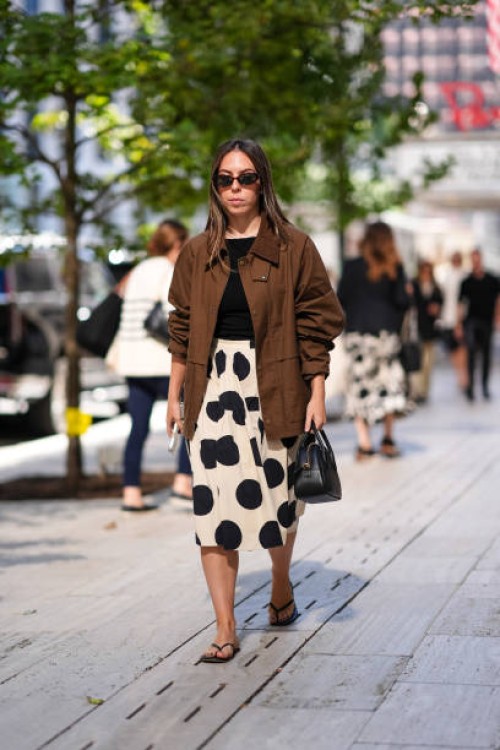 Guest wears black sunglasses, black shirt, white black polka dotted pattern/print pleated midi skirt, dark brown oversized jacket, shiny black...