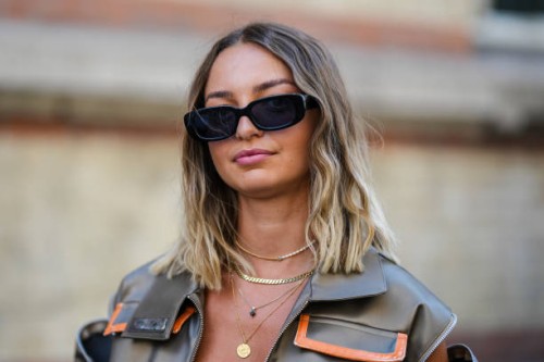 Guest wears black sunglasses, gold chain pendant necklaces, a dark gold shiny sleeveless / zipper pockets / cargo oversized jacket, during the...