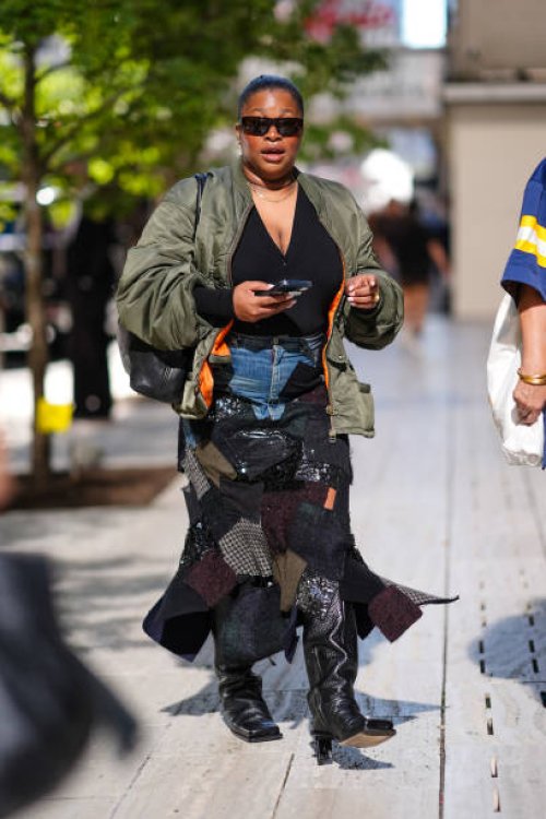Guest wears black sunglasses, gold necklace, black v-neck shirt, dark green bomber jacket, navy blue denim jean mixed patch midi skirt, shiny black...