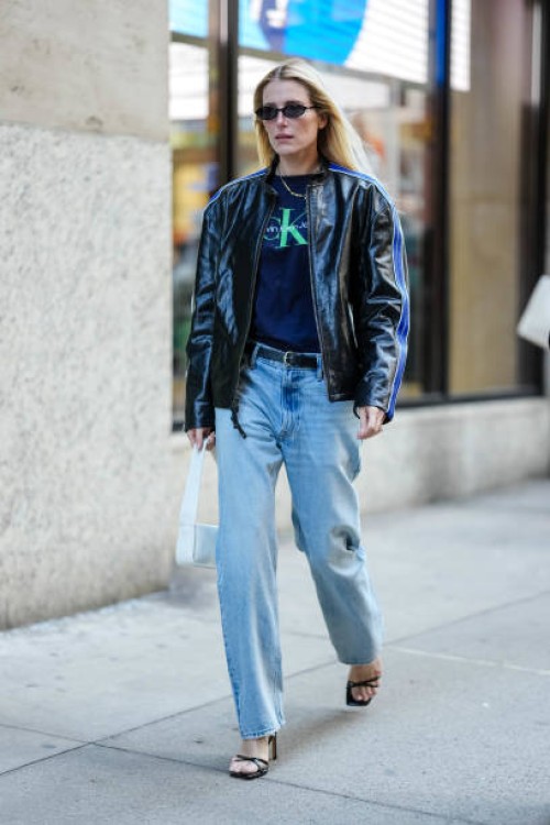 Guest wears black sunglasses, gold necklace, dark navy blue bright green Calvin Klien Jeans print t-shirt, shiny black dark blue lined unzipped biker...