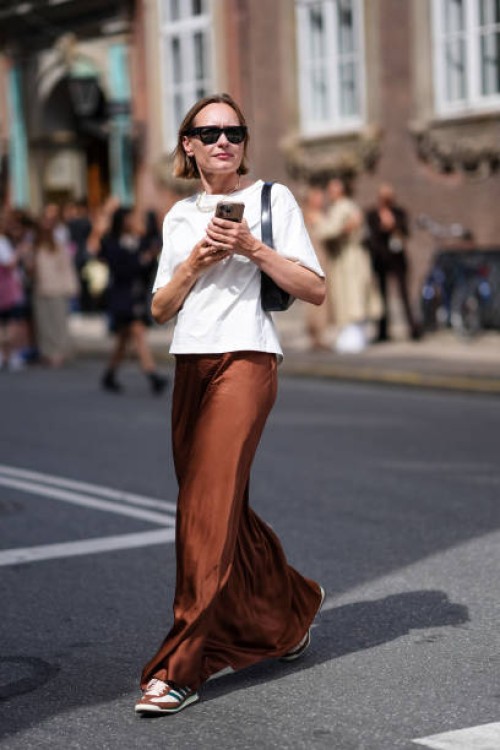 Guest wears black sunglasses, gold necklace, white short sleeve shirt, black leather bag, shiny dark brown silk maxi skirt, White brown Adidas...