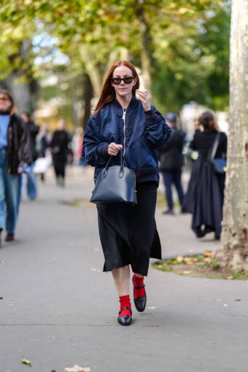 Guest wears black sunglasses, navy blue bomber jacket, shiny dark navy blue leather bag, black satin midi skirt, bright red socks, shiny black...
