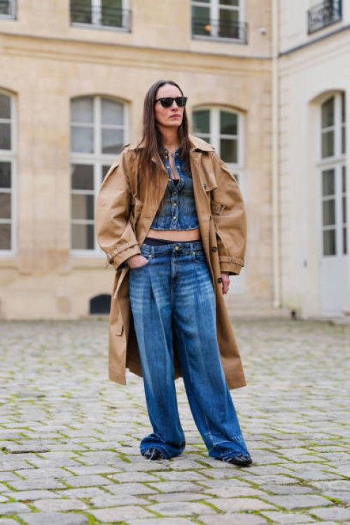 Guest wears black sunglasses, navy blue buttoned up denim jean jacket, light brown trench coat, navy blue washed loose denim jean pants, outside...
