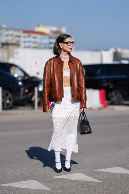 Guest wears black sunglasses, pearl earrings, pearl layered choker necklace, white bralette, shiny dark brown unzipped leather jacket, white high...