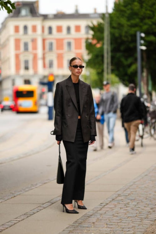 Guest wears black sunglasses, silver earrings, a black cropped top, a black oversized blazer jacket, a black high waist long skirt, a black shiny...