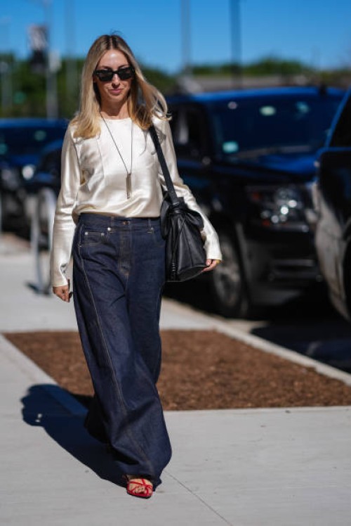 Guest wears black sunglasses, silver necklace with pendant, cream lustrous silky long sleeve shirt, dark navy blue denim loose pants, bright red...