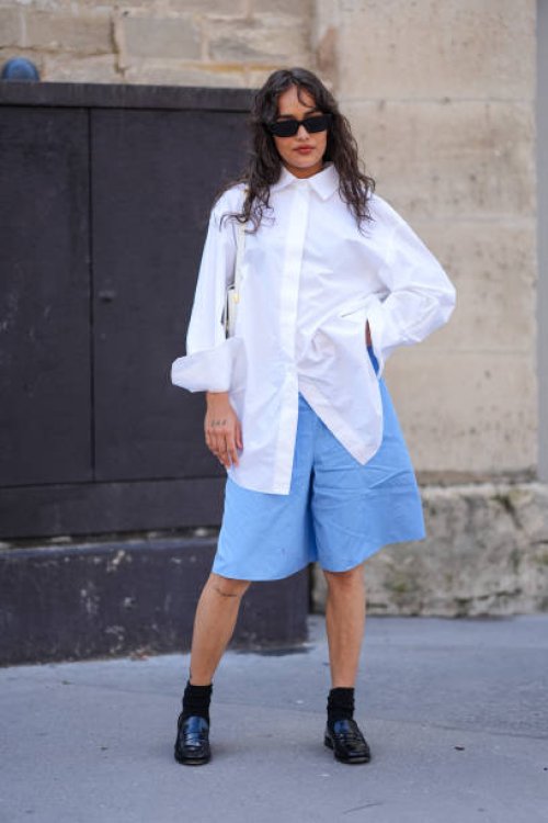 Guest wears black sunglasses, white oversized button up long sleeve shirt, white leather bag, light blue shorts, black socks, shiny black loafer...