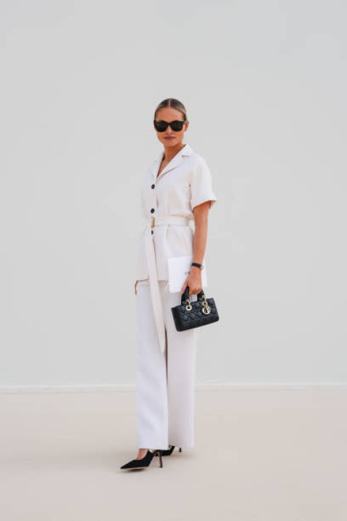 Guest wears black sunglasses, white short sleeve belted buttoned up blazer jacket, white straight trouser pants, shiny black quilted Dior Lady D-Joy...