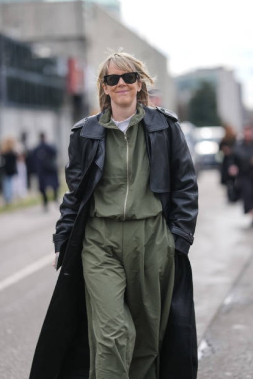 Guest wears black sunglasses, white t-shirt, dark olive green zipped bomber jacket, dark olive green loose trouser pants, shiny black leather trench...