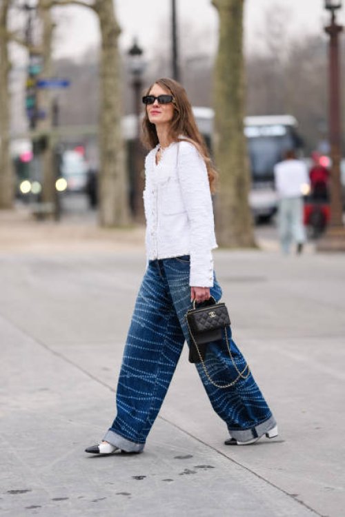 Guest wears black sunglasses, white tweed pearl detail buttoned Chanel blazer jacket, navy blue white pearl pattern/print baggy Chanel denim jean...