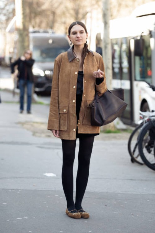 Guest wears black tights, brown suede jacket, dark brown bag and suede flats outside the Christopher Esber fashion show during the Womenswear...
