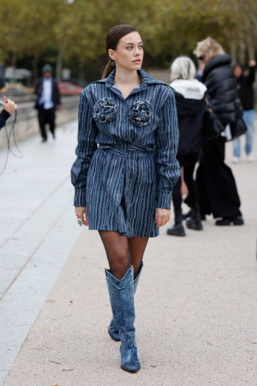 Guest wears blue denim striped shirt dress with flowers detail, black sheer tights, denim cowboy boots, outside Balenciaga, during Womenswear...
