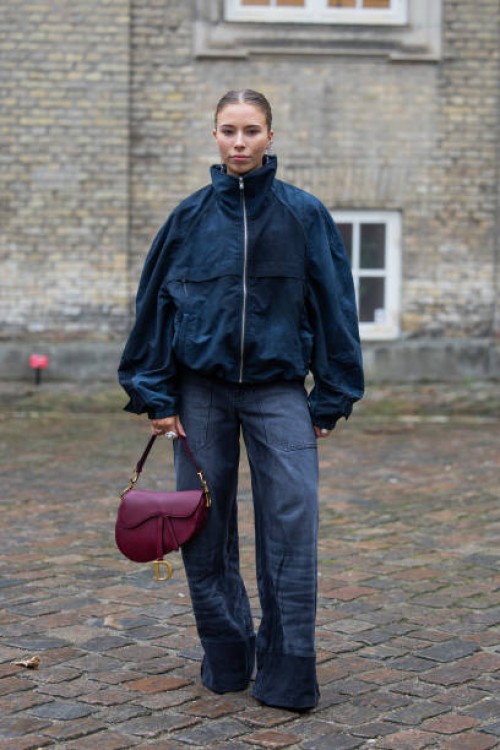 Guest wears blue jacket, Dior saddle bag, wrapped wide leg denim jeans outside TG Botanical during the Copenhagen Fashion Week Autumn/Winter 2025 on...
