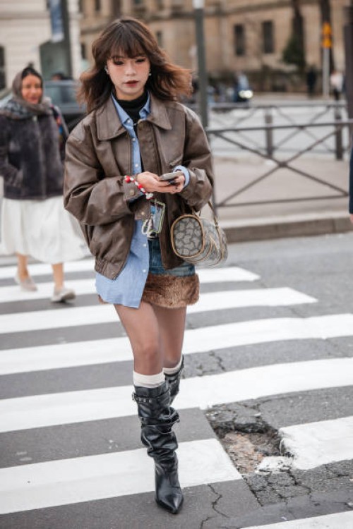 Guest wears blue shirt, brown leather jacket, denim mini skirt with fur detail, black boots, brown bag, outside Miu Miu, during the Womenswear...