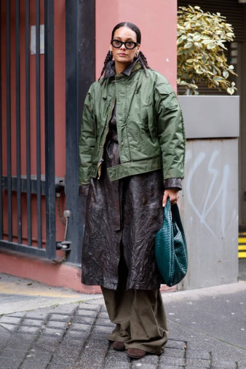 Guest wears brown leather trench coat, olive green pants, brown boots, olive green jacket, green Bottega Veneta bag, black Sunnei sunglasses, outside...