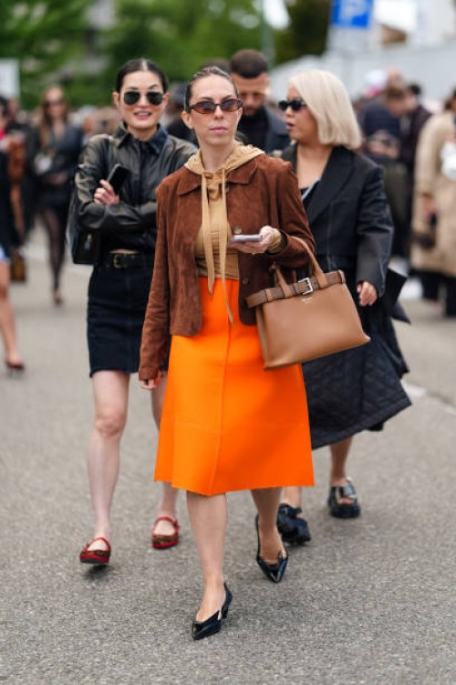 Guest wears brown sunglasses, a brown suede jacket, a light brown hoodie, a brown Prada leather handbag, a long flashy orange skirt, black pointed...