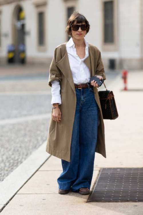 Guest wears brown sunglasses, brown leather loafers, a brown leather belt, a white shirt, a beige trench, blue wide leg jeans and a black and brown...