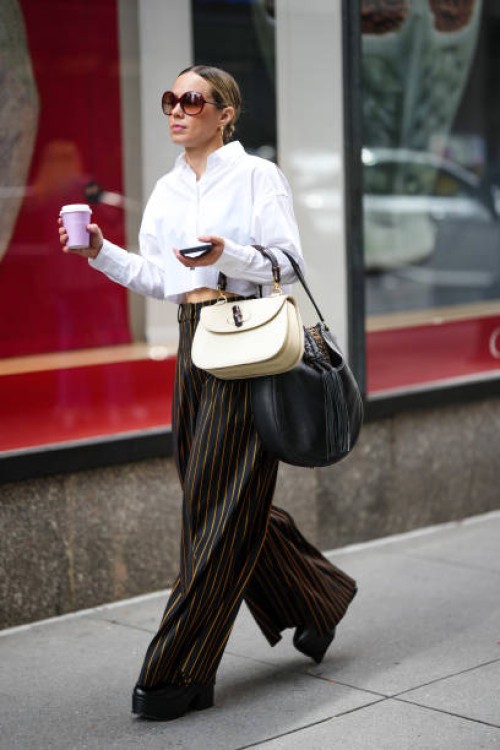 Guest wears brown sunglasses, gold earrings, a silver large chain necklace, a white cropped shirt, black with small gold striped print pattern large...