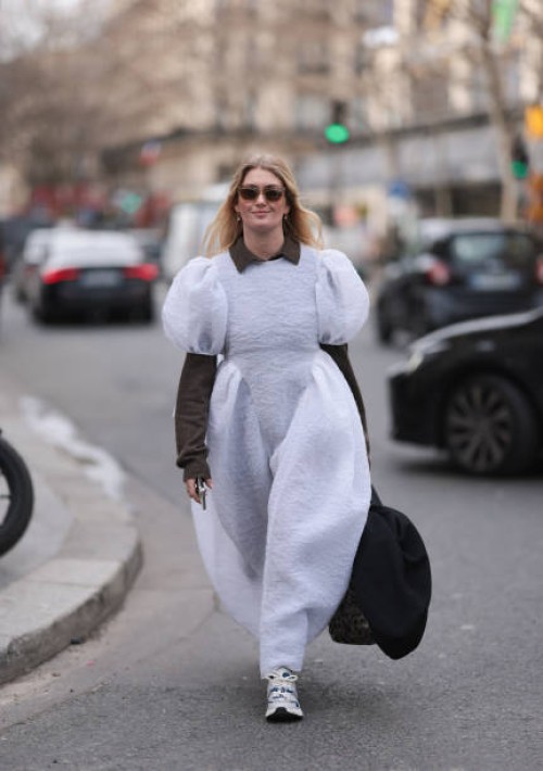 Guest wears brown sweater under white puffy maxi dress, sneakers, tote bag and black sunglasses and white sneaker outside Dries Van Noten show during...