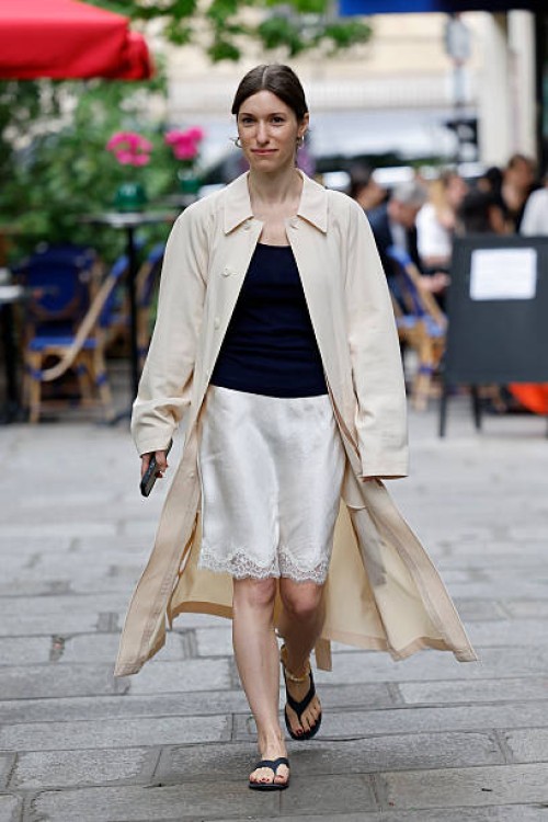 Guest wears dark blue top, white silk skirt with lace, beige trench coat, outside Lemaire, during the Menswear Spring/Summer 2026 show as part of...