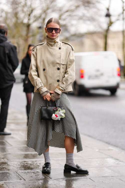 Guest wears dark brown sunglasses, gold earrings, light brown high neck double breasted trench jacket, gold bracelets, gold rings, white black...