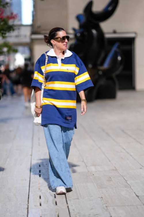 Guest wears dark brown sunglasses, white navy blue yellow striped short sleeve oversized shirt, white leather bag, a gold bracelet, a watch, gold...