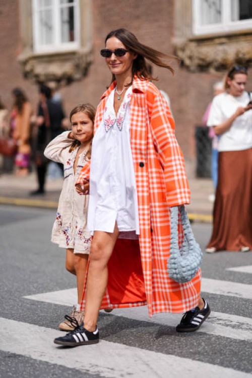 Guest wears dark brown sunglasses, white necklace, bright orange and white checkered pattern trench coat, white mini dress with large collar and...