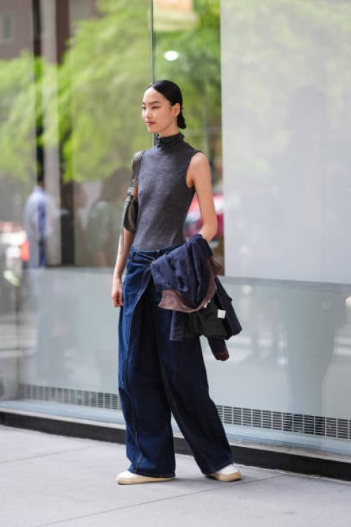 Guest wears dark gray sleeveless turtleneck shirt, dark gray belted bag, navy blue denim jean pants, black jacket in hand, white sneaker shoes,...