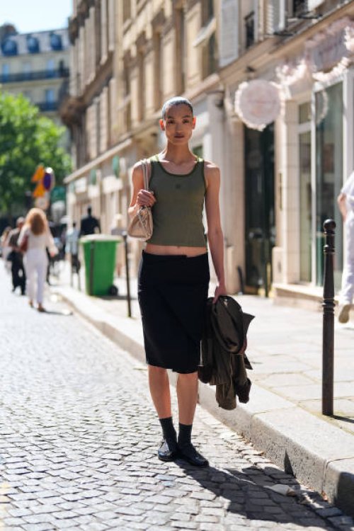 Guest wears dark green tank top, beige leather bag, black low-rise tailored pencil skirt, black socks, shiny black loafer moccasin leather shoes,...