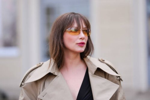 Guest wears dark orange sunglasses, black deep v-neck shirt, light brown trench coat, outside Tod’s, during the Milan Fashion week Women's...