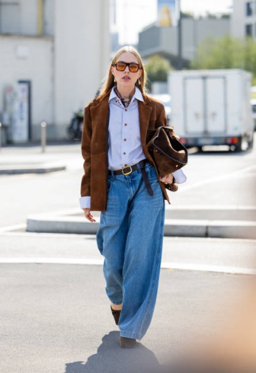 Guest wears denim wide leg jeans, brown jacket, bag, blue button shirt outside Prada during the Milan Fashion Week Menswear Spring/Summer 2025 on...